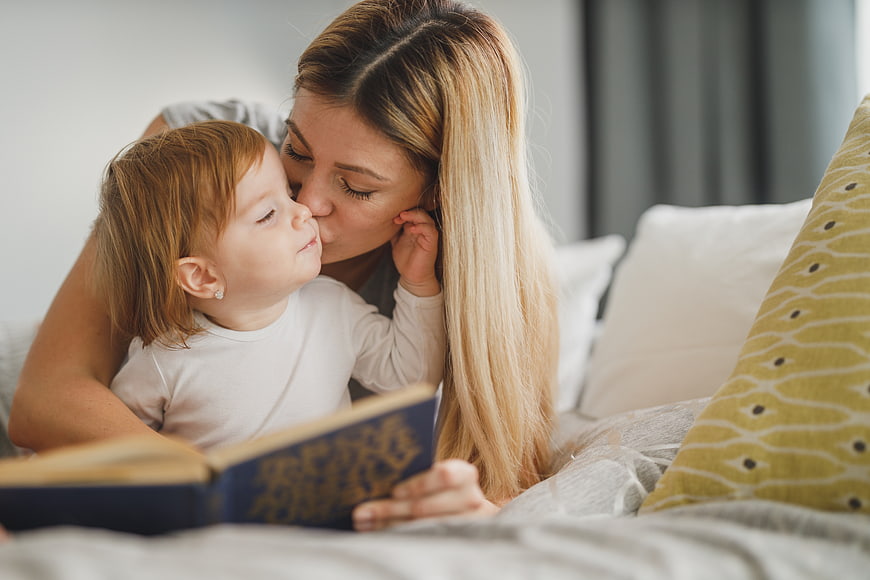 Mother kissing toddler daughter while reading book in bed