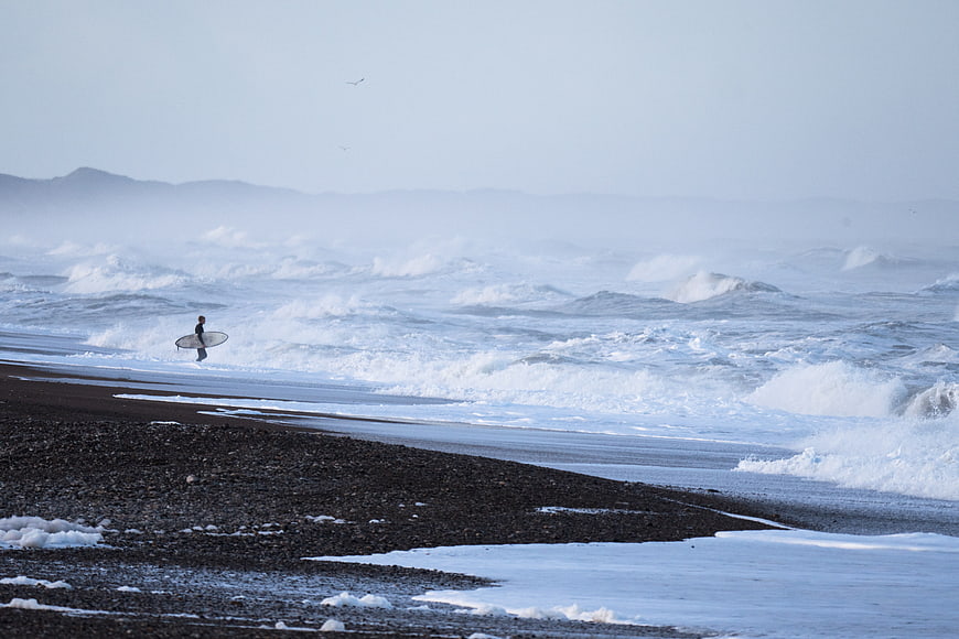 Lone Surfer Walking on Black Sand Beach with Rough Waves
