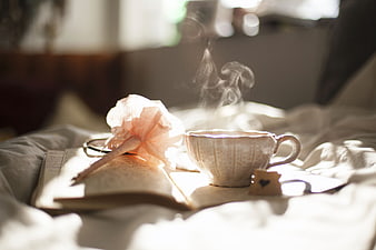 Steaming cup of tea on bed with open book and flower