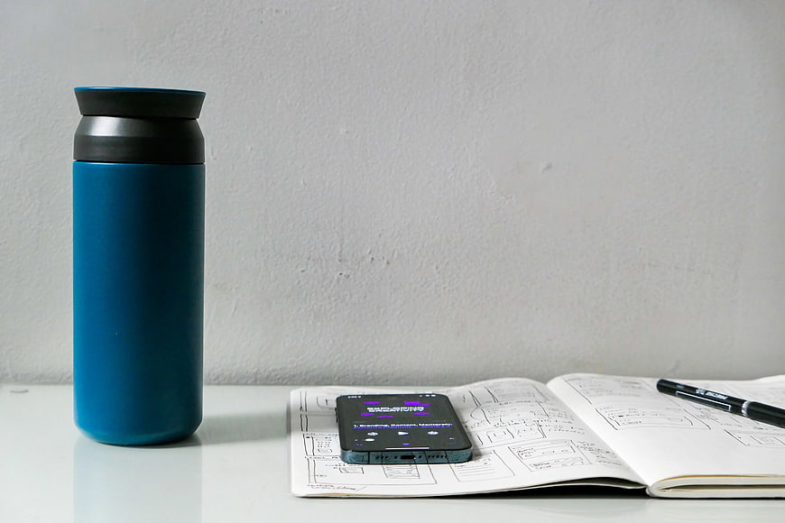 Blue Thermos, Smartphone, and Sketchbook on White Desk