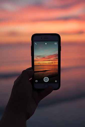 Hand holding smartphone capturing sunset over ocean
