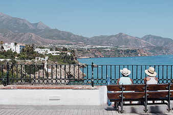 Two women in hats sitting on a bench overlooking the sea and mountains