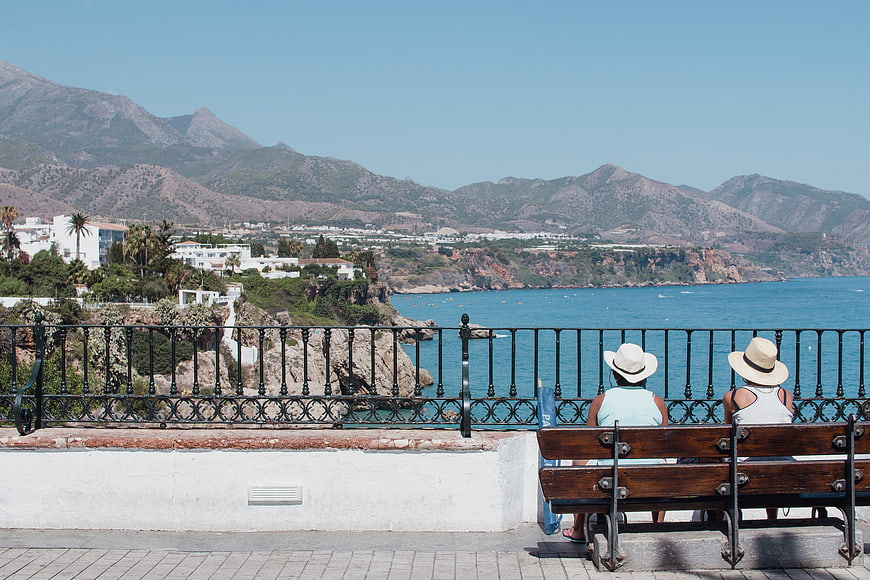 Two women in hats sitting on a bench overlooking the sea and mountains