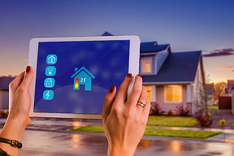 Hands holding tablet with smart home app interface in front of suburban house