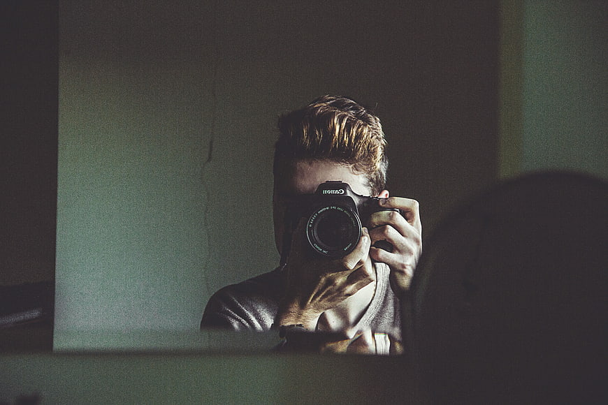Man taking mirror selfie with DSLR camera in dim room