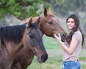 Young woman smiling while petting two brown horses in a green pasture