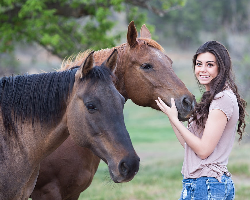 Young woman smiling while petting two brown horses in a green pasture