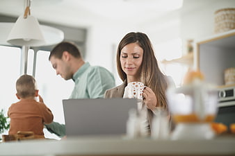 Mother working on laptop with coffee while family has breakfast