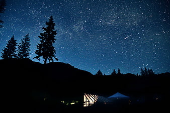 Starry Night Sky Over Mountain Silhouette with Glowing Cabin