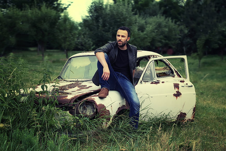 Man Sitting on Abandoned Rusty Car in Overgrown Field