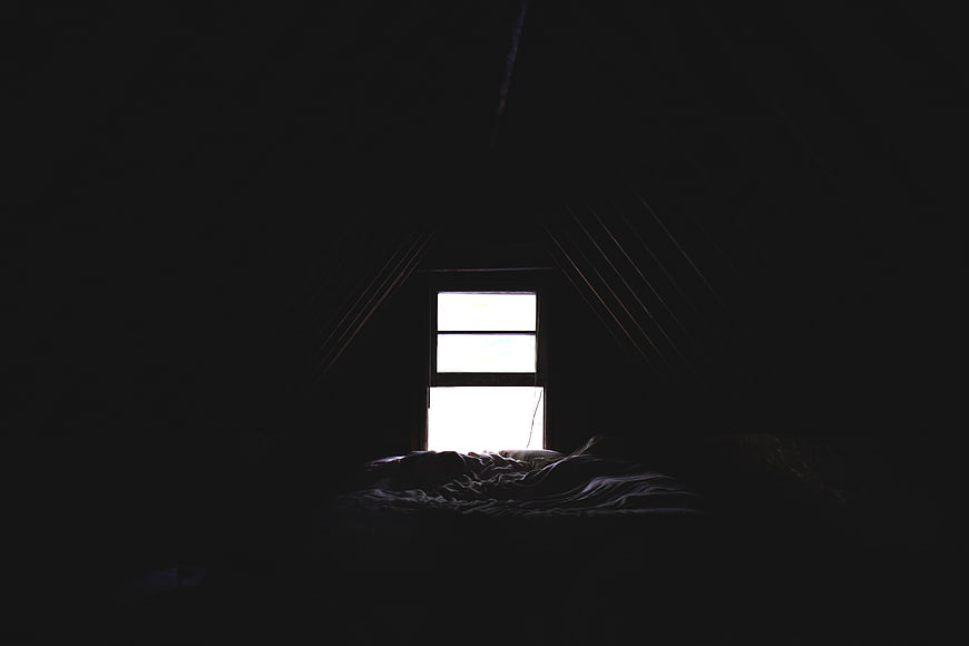 Dark attic bedroom with bright window and unmade bed