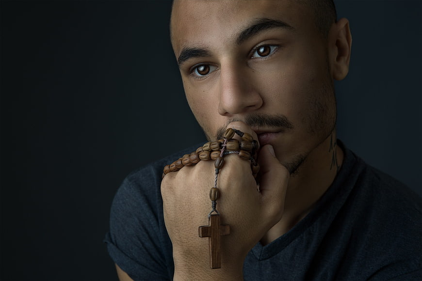 Young man praying with wooden rosary and cross against dark background