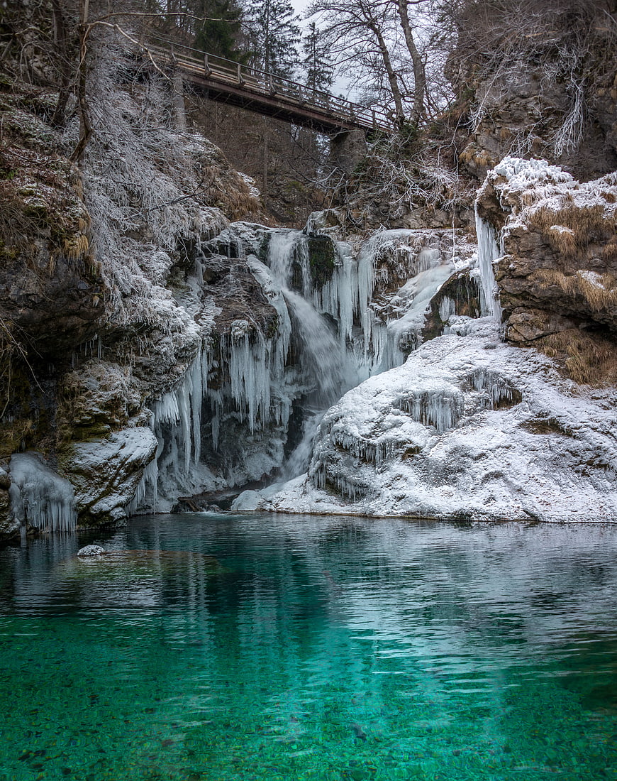 Frozen waterfall cascading into turquoise pool with wooden bridge