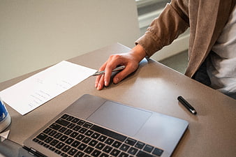 Hand holding pen over laptop and paper on desk