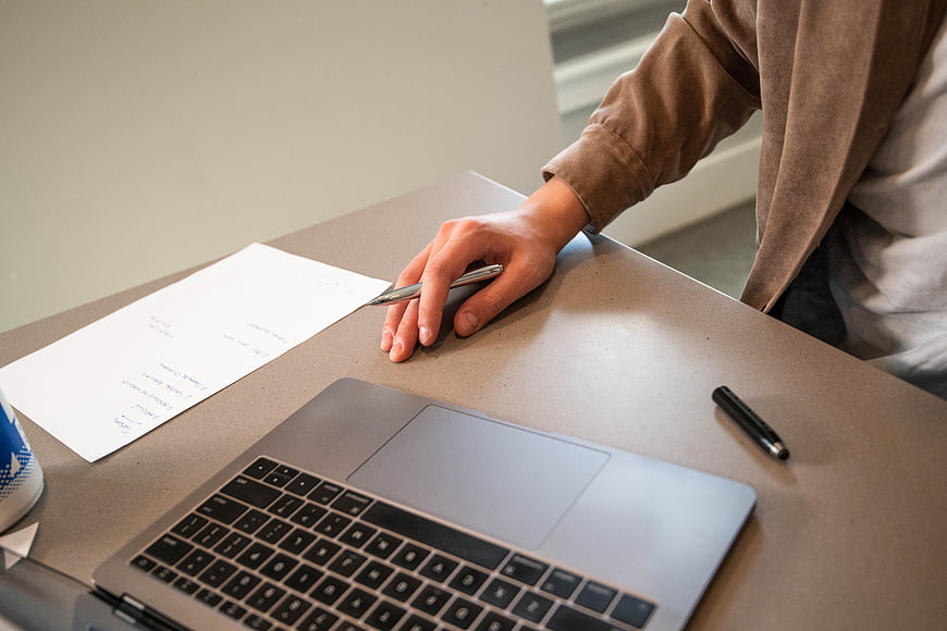 Hand holding pen over laptop and paper on desk