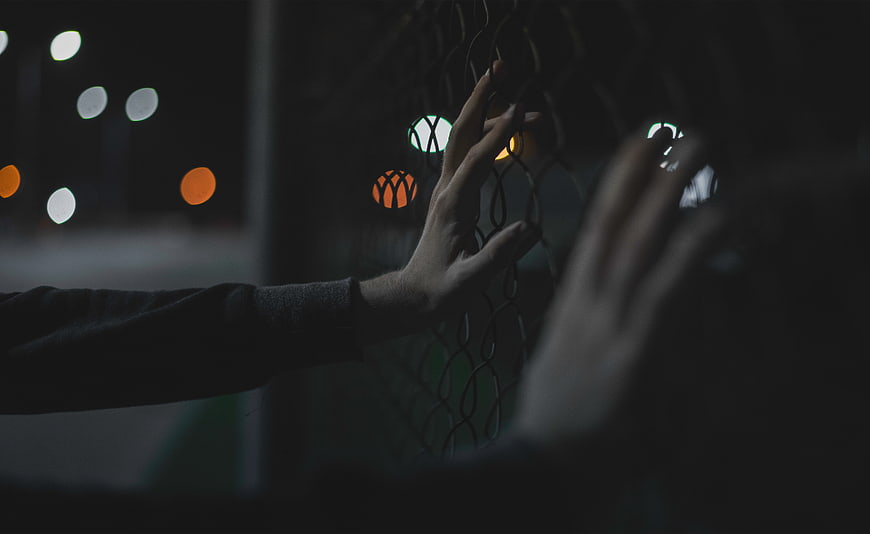 Hands touching chain link fence at night with bokeh lights