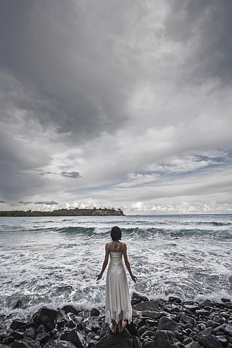 Woman in White Dress Standing on Rocky Beach Facing Stormy Ocean