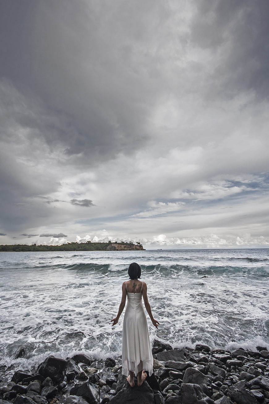 Woman in White Dress Standing on Rocky Beach Facing Stormy Ocean