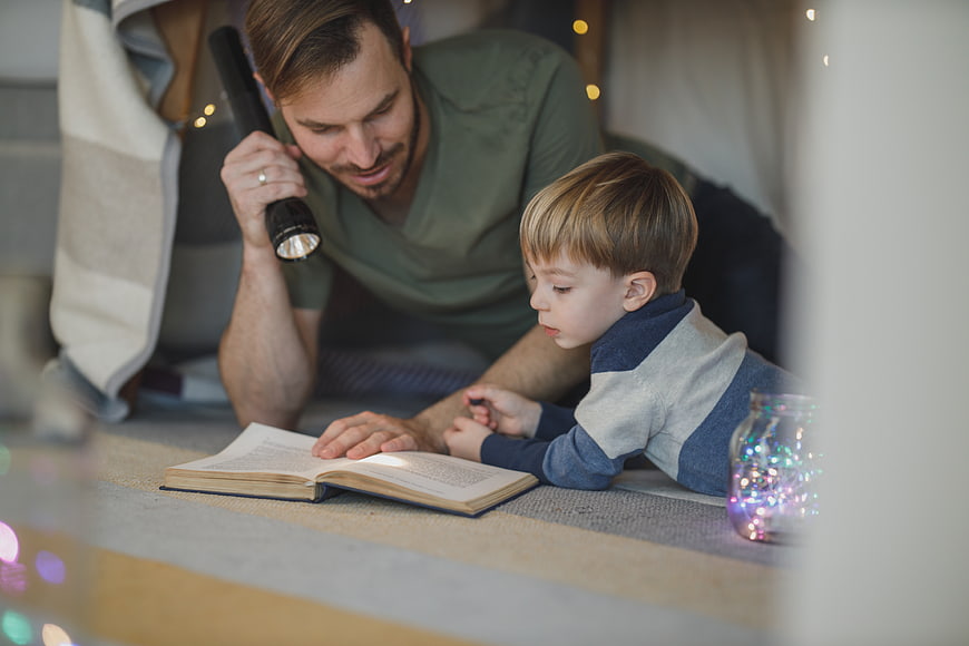 Father and son reading book with flashlight in cozy indoor tent