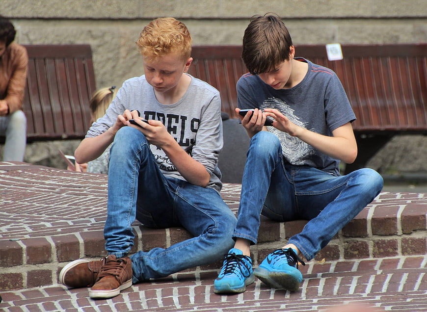 Two teenage boys playing mobile games on smartphones sitting on brick steps