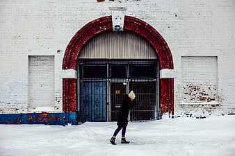 Woman walking in snow past white brick building with red arch