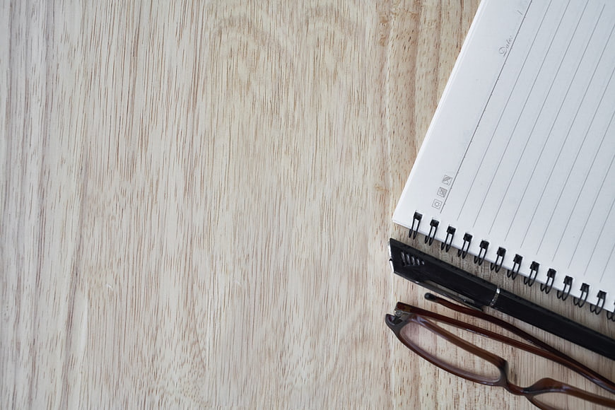 Flat lay of notebook, pen, and glasses on light wood table