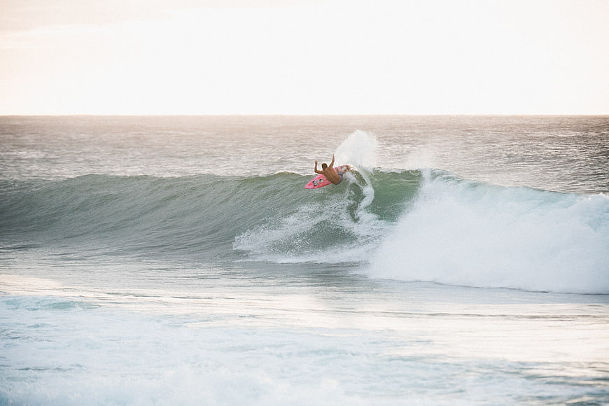 Surfer riding a wave on a pink surfboard at sunset