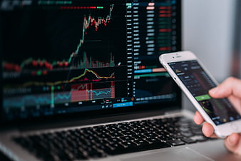 Hand Holding Smartphone With Trading App In Front Of Laptop Stock Charts