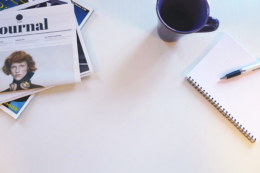 Flat lay of magazine, coffee mug, and notebook on white desk