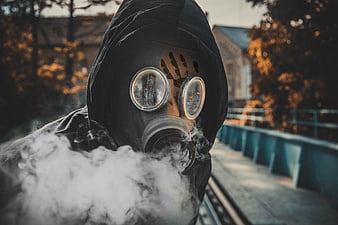 Person in black hood and gas mask exhaling smoke on a bridge