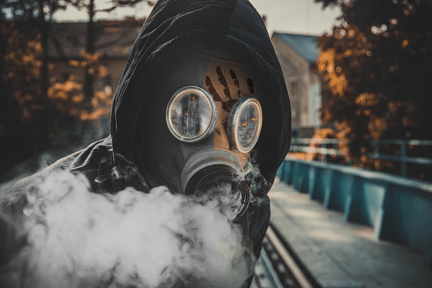 Person in black hood and gas mask exhaling smoke on a bridge