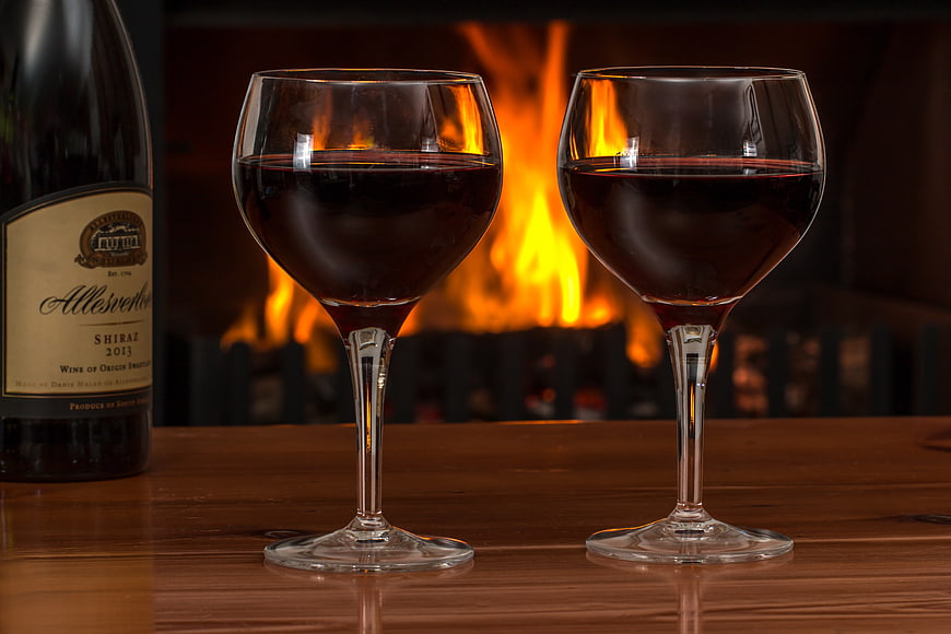 Two glasses of red wine on a wooden table in front of a fireplace
