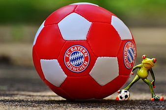 Toy frog kicking miniature soccer ball next to large red Bayern Munich ball
