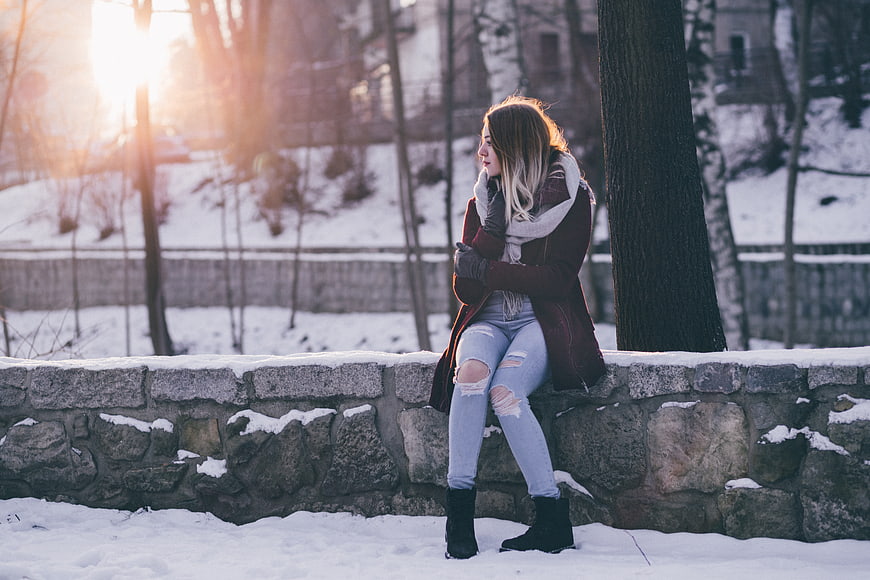 Woman in winter coat sitting on stone wall in snowy park