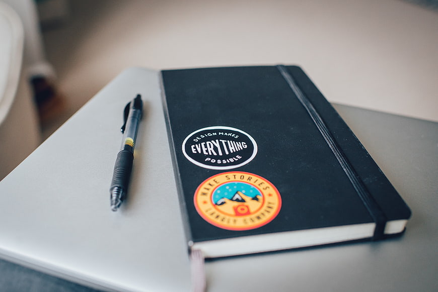 Black Notebook with Stickers and Pen on Laptop