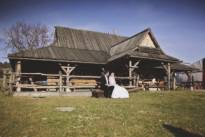 Wedding couple sitting on rustic wooden bench in front of traditional house