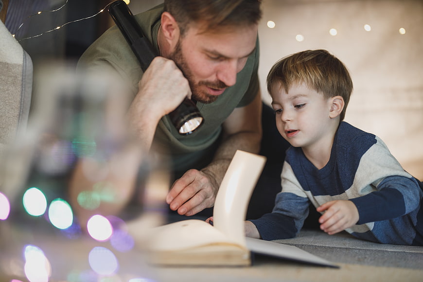 Father and son reading book with flashlight indoors