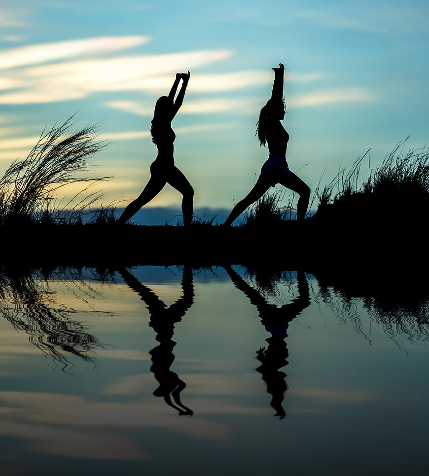 Silhouettes of Two Women Doing Yoga by Water at Sunset