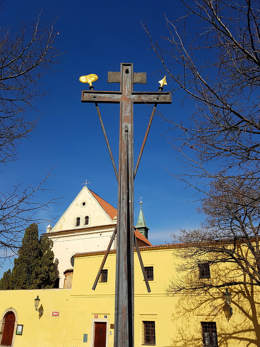 Rusty metal cross with golden flames against blue sky and yellow church
