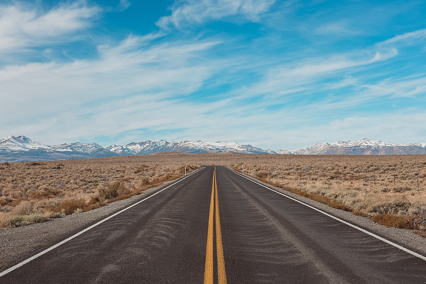 Straight desert highway leading to snow-capped mountains under blue sky