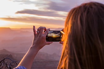 Woman photographing sunset over canyon landscape with smartphone
