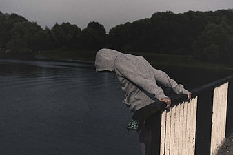 Anonymous hooded figure leaning over bridge railing in despair