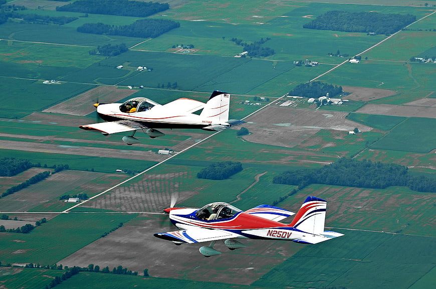Two Light Sport Aircraft Flying In Formation Over Green Farmland