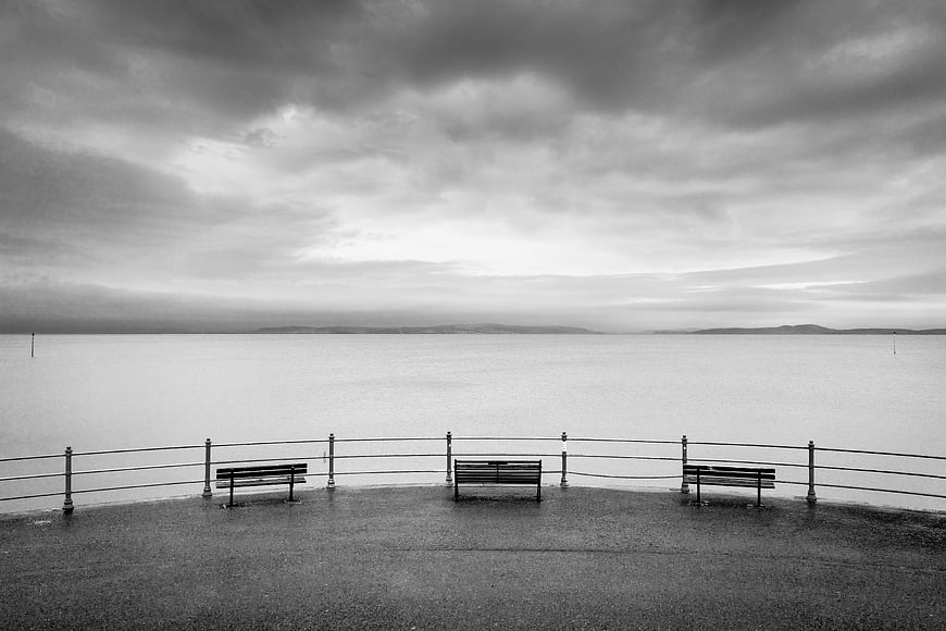 Black and white photo of empty benches on a pier overlooking the sea