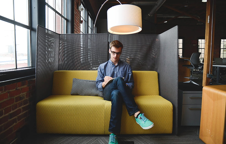Young man writing on yellow sofa in modern office