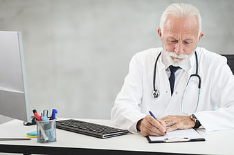 Senior male doctor writing on clipboard at office desk