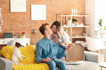 Happy young couple kissing while eating pizza on sofa with dog