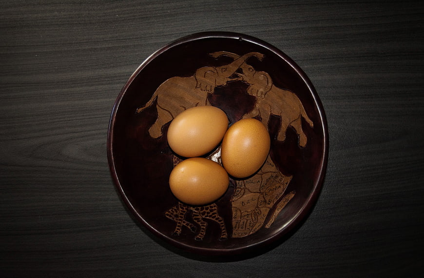 Three brown eggs in a carved wooden bowl on dark table
