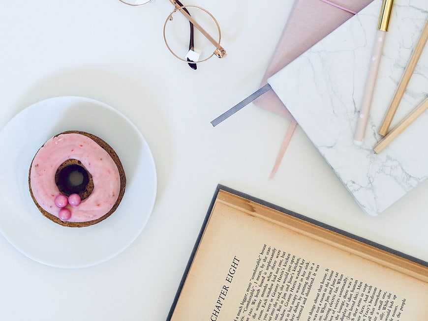 Flat lay of pink donut, book, and stationery on white desk