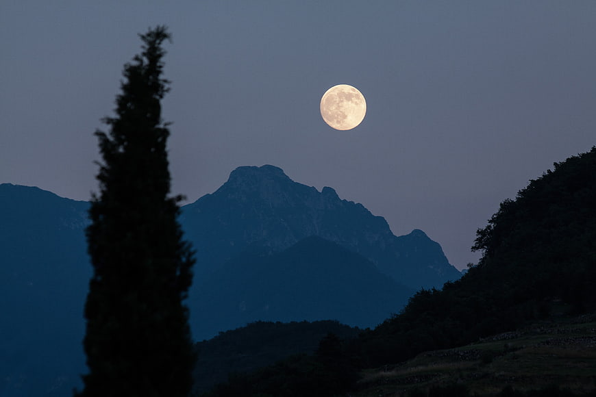 Full moon rising over dark mountain silhouette with cypress tree